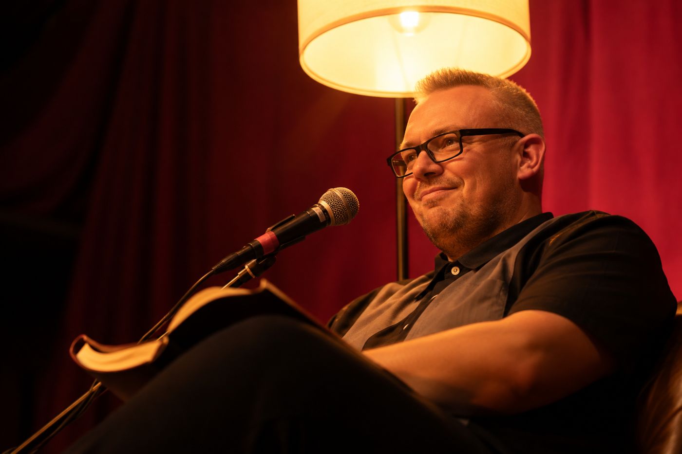 Dr. Benjamin Förster liest im Sessel unter einer Stehlampe, vor ihm ein Mikrofon